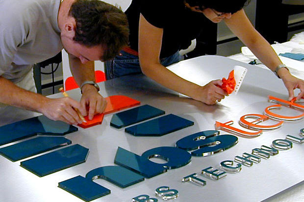 Signage installers working on vinyl for dimensional acrylic letters on brushed stainless steel metal background