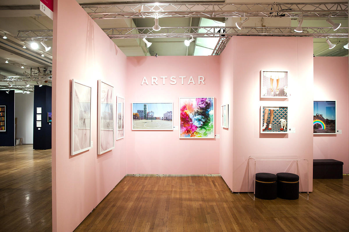 An Art Show booth with pink walls, raised 3D letters that say ArtStar and colorful paintings in white frames.
