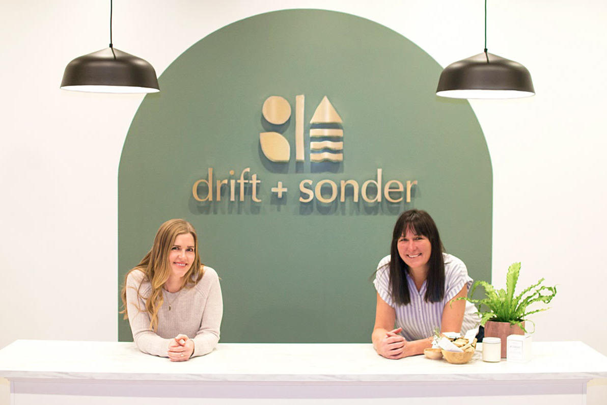Two women standing behind the front counter of Drift and Sonder Lifestyle Shop Ltd. in Fernie, BC, with a custom minimalist 3D sign featuring the shop’s logo and name mounted on a green accent wall, created by ArtSigns.