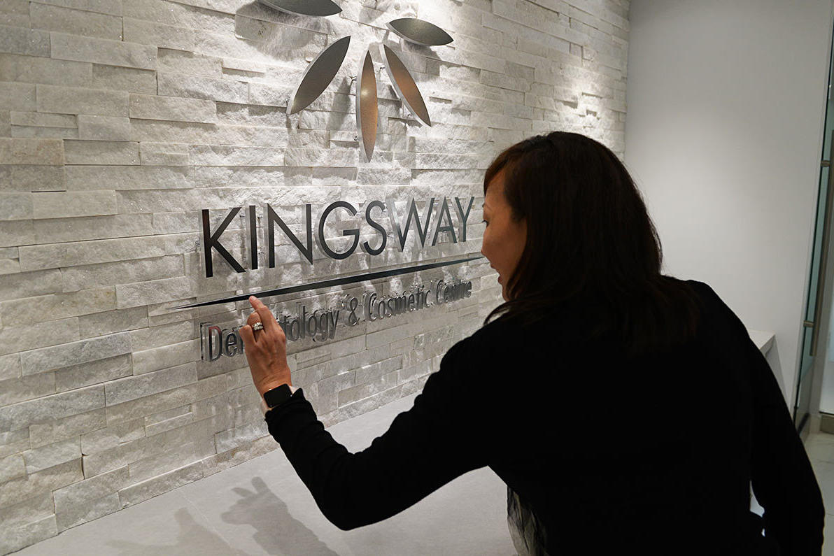 A woman closely examining a high-quality, acrylic and metal 3D sign for Kingsway Dermatology & Cosmetic Centre, mounted on a textured stone wall. The polished finish and precise craftsmanship highlight the superior materials used in the sign.