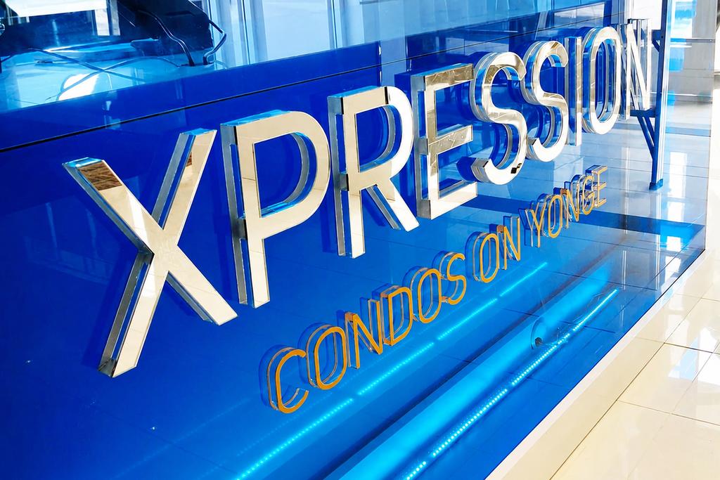 A close-up view of the XPRESSION Condos on Yonge sales centre sign featuring raised chrome letters mounted on a vivid blue acrylic panel with gold “Condos on Yonge” dimensional lettering
