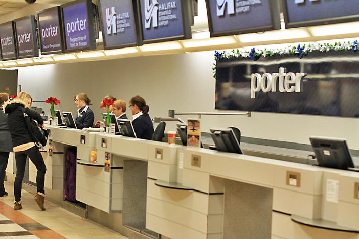 Porter Airlines sign mounted on a check-in counter wall at Halifax Stanfield International Airport. The area features multiple check-in kiosks, staff assisting travelers, and overhead digital displays showing the Porter logo.