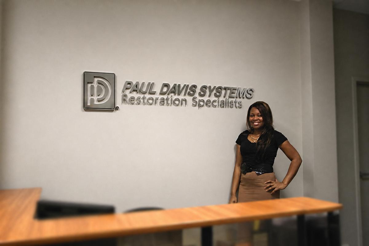 Staff member beside a dimensional Paul Davis reception wall sign behind a desk